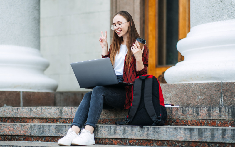 A young woman with glasses sits on university steps with a laptop and coffee, making a peace sign. She has a backpack beside her and looks cheerful while studying.