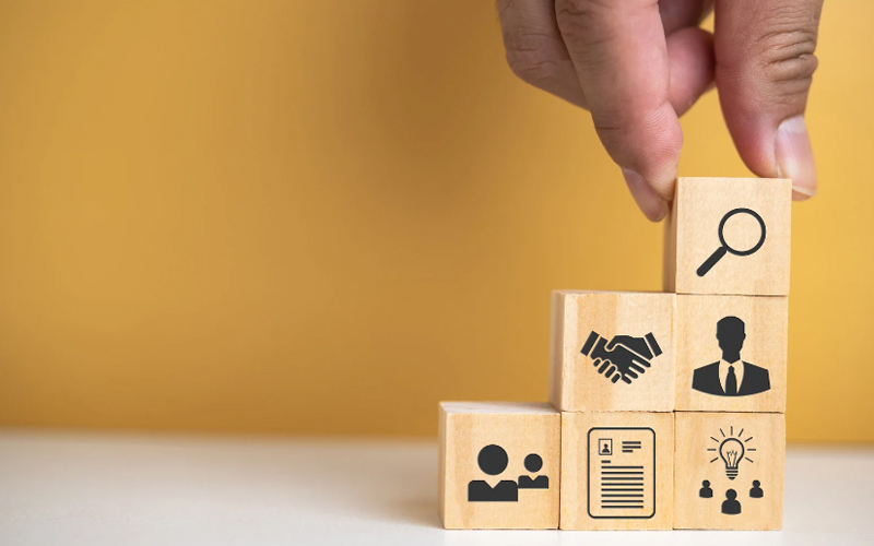 A hand arranges wooden blocks with icons representing career elements: job search, networking, resume, teamwork, and ideas.