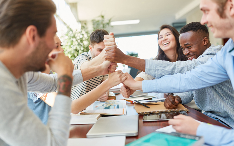A group of diverse students sitting around a table, smiling and giving each other high-fives in a celebratory gesture.